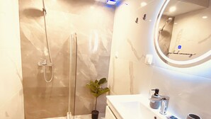 Une salle de bain moderne avec une douche à parois de verre, un grand miroir rond avec éclairage intégré et un lavabo blanc doté d’un distributeur de savon.