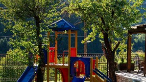 Un'area giochi con scivoli e strutture per arrampicata, circondata da alberi e con montagne sullo sfondo.
