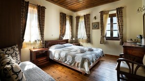 Ein Schlafzimmer mit Holzbalkendecke, einem Bett mit gemustertem Überwurf, einem Nachttisch, einem Schreibtisch und einem Sessel.