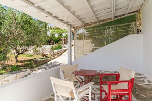 A covered outdoor seating area with two chairs and a table, overlooking a garden with trees and a stone wall.