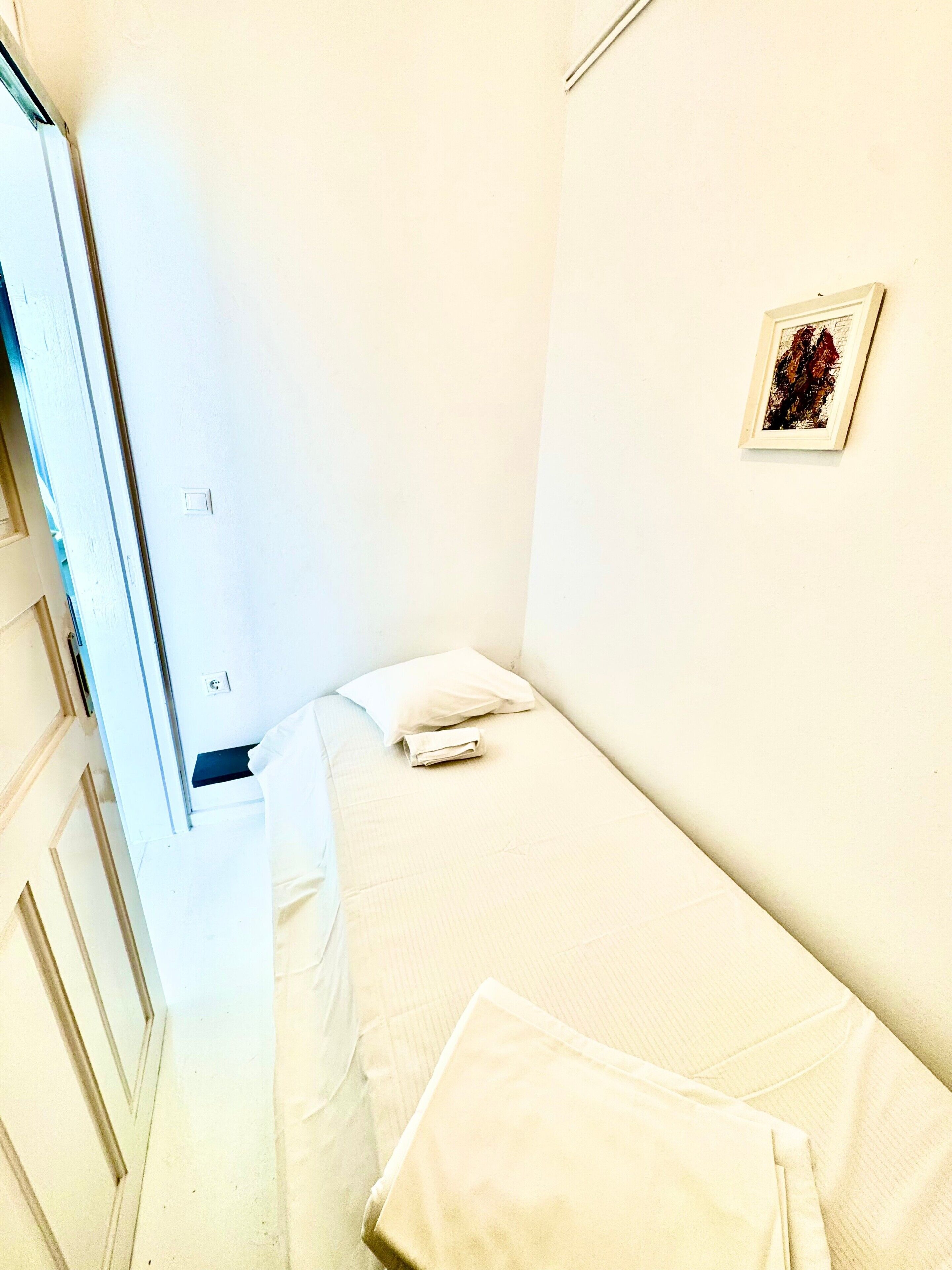 A small, single-bed room with a white bed, a pillow, a folded towel, a framed picture on the wall, and a door with a handle.