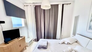 A hotel room with a bed, a television, a bedside table, a window with curtains, and a view of the sea. - Apartment 'Lefkopetra' with Shared Terrace, Wi-Fi and Air Conditioning (Tinos)