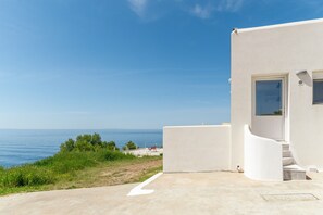 Un bâtiment blanc avec un escalier menant à une porte, une vue dégagée sur la mer et une zone herbeuse avec un sentier.