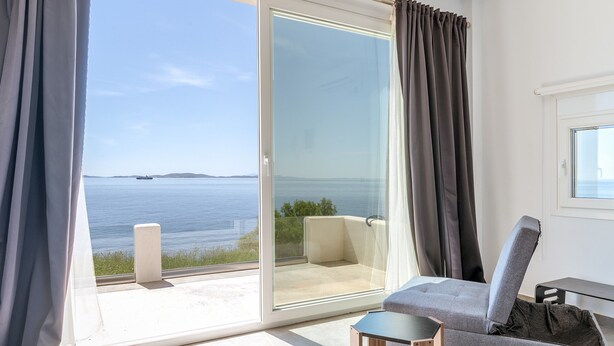 A room with a sofa, a small table, and a view of the sea through a glass door. - Studio 'Theasea' with Private Garden, Wi-Fi and Air Conditioning (Tinos)