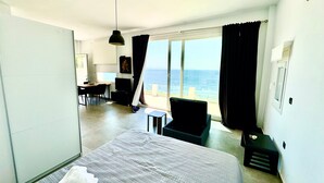 A hotel room with a bed, a desk, a chair, a TV, and a large window with a view of the sea. - Studio 'Theasea' with Private Garden, Wi-Fi and Air Conditioning (Tinos)
