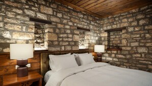 A bedroom with a stone wall, wooden headboard, two bedside tables with lamps, and a bed with white bedding. - Deluxe 'Dryopi' Apartment with Mountain View, Private Terrace & Wi-Fi (Karpenision)