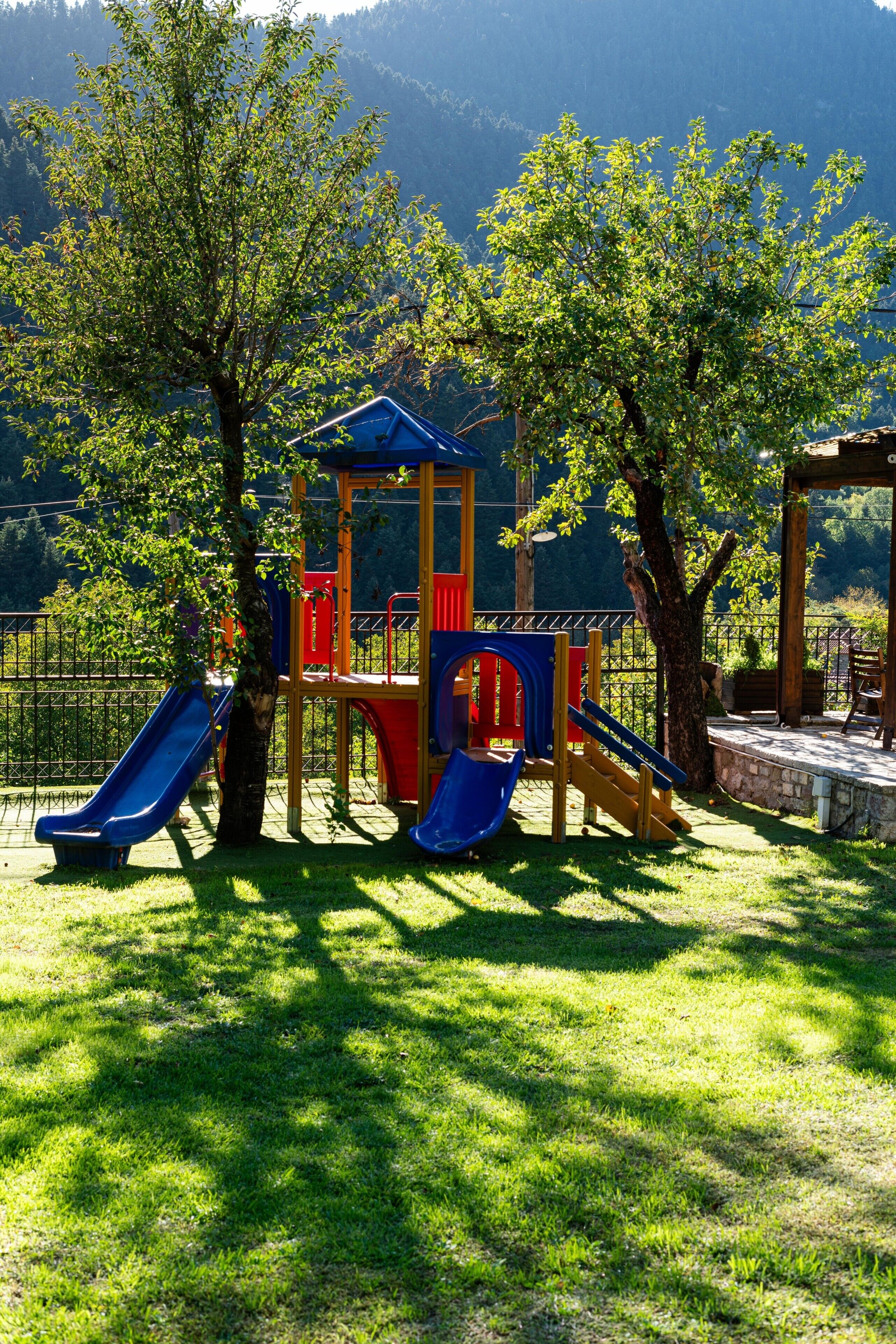 A playground with slides and climbing structures, surrounded by trees and a mountainous backdrop.