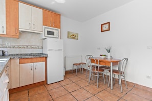 A kitchen with wooden cabinets, a white refrigerator, a microwave, a dining table with four chairs, and a clock on the wall.