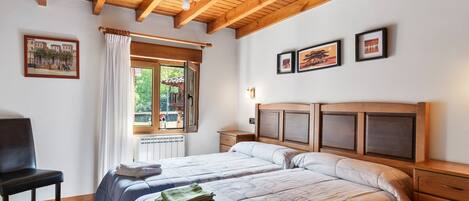 A bedroom with a wooden ceiling, a bed with a headboard, a chair, a window with curtains, and framed pictures on the wall.