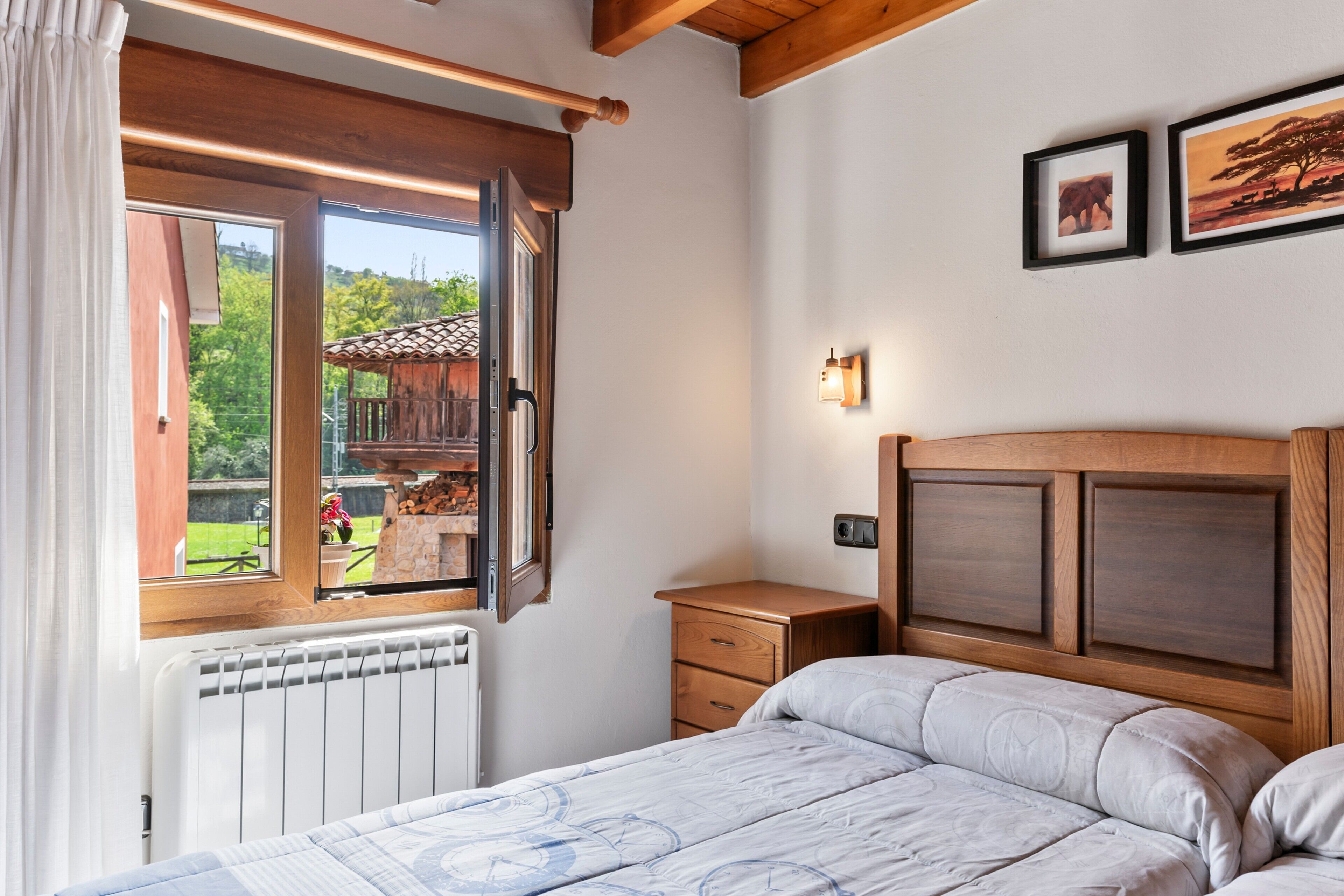 A bedroom with a wooden headboard, a bed with a patterned bedspread, a nightstand, a dresser, a window with an open shutter, and a view of a wooden structure outside.