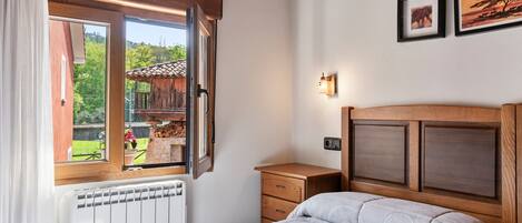A bedroom with a wooden headboard, a bed with a patterned bedspread, a nightstand, a dresser, a window with an open shutter, and a view of a wooden structure outside.
