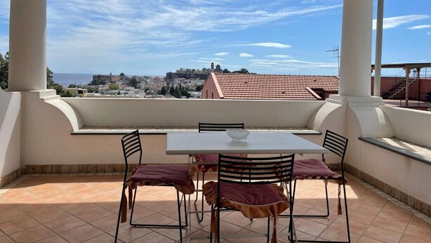 Un patio al aire libre con una mesa y sillas, vista a un pueblo y al mar, bajo un cielo despejado.