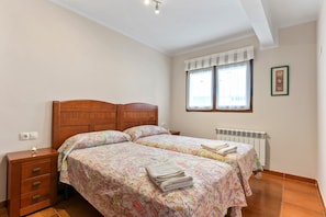 A bedroom with two beds, a nightstand, a radiator, and a window with curtains.