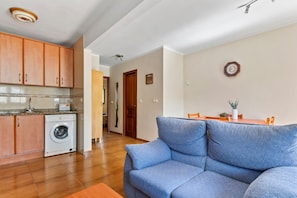 Ein Wohnzimmer mit blauem Sofa, einer Waschmaschine, Holzsärchen und einem Essbereich mit Tisch und Stühlen.