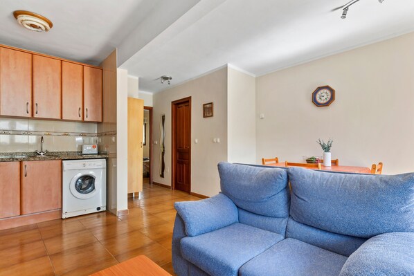 Una sala de estar con un sofá azul, una lavadora, armarios de madera y un comedor con mesa y sillas.