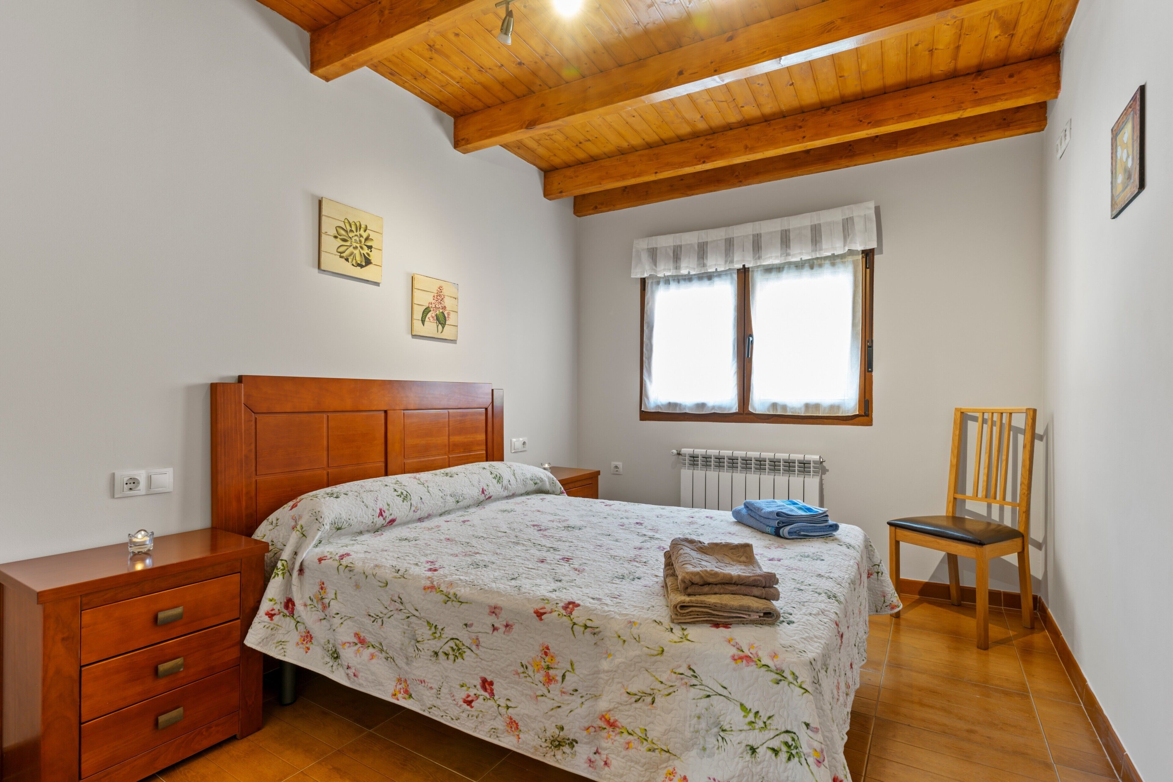 Un dormitorio con cama de madera, mesita de noche, silla, radiador y ventana con cortinas.