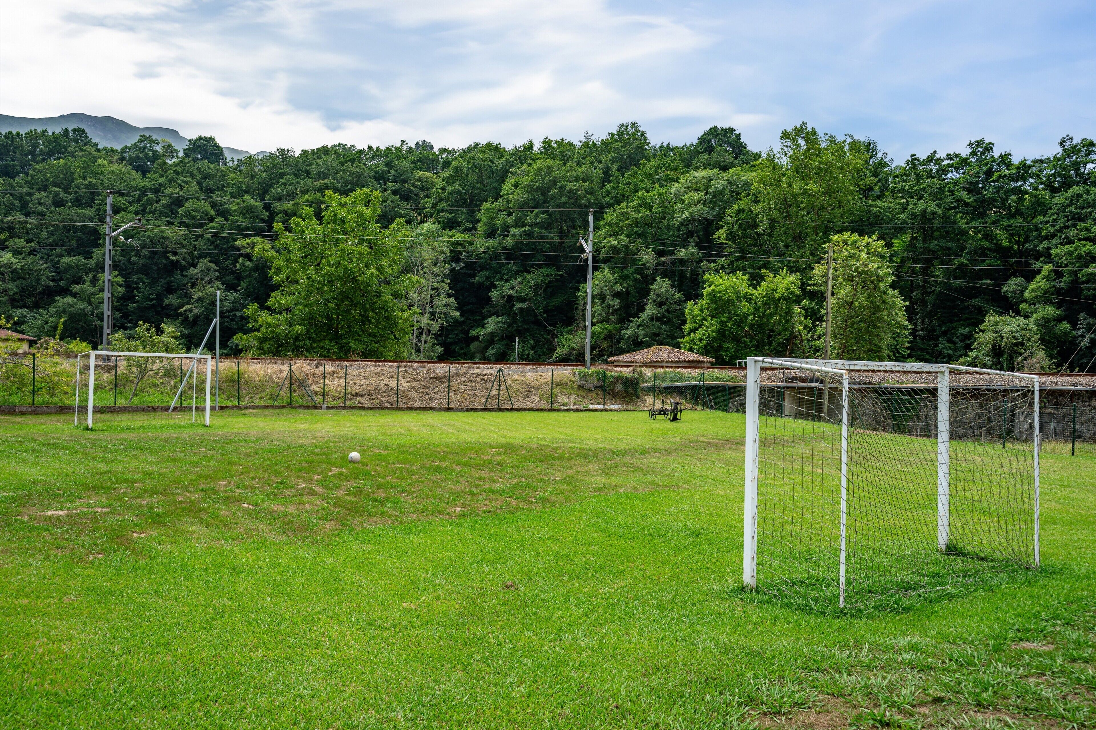 A soccer field with two goals, surrounded by a fence, and a forested hillside in the background.