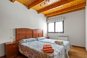 Une chambre avec un plafond en bois, un lit double avec une literie à motifs floraux, une table de chevet et une fenêtre agrémentée d’une lambrequin blanche.
