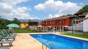 A swimming pool with lounge chairs and umbrellas, surrounded by buildings and a mountainous backdrop. - Casa rural 'El Trasgu' con vistas a la montaña, piscina y barbacoa (Unknown)