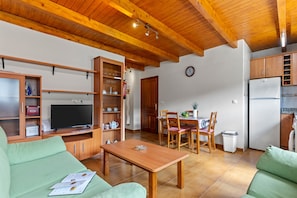 Una sala de estar con televisor, muebles de madera, un comedor con sillas y una mesa, y una cocina con refrigerador.