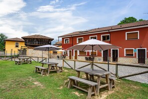 Outdoor wooden picnic tables with umbrellas, a stone pathway, and a red building with windows and a door. - Casa rural 'El Trasgu' con vistas a la montaña, piscina y barbacoa (Unknown)