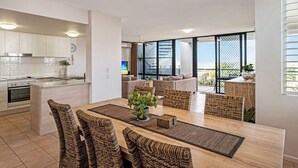 Une cuisine moderne avec des meubles blancs, un îlot central et un coin repas comprenant des chaises en osier et une table en bois.