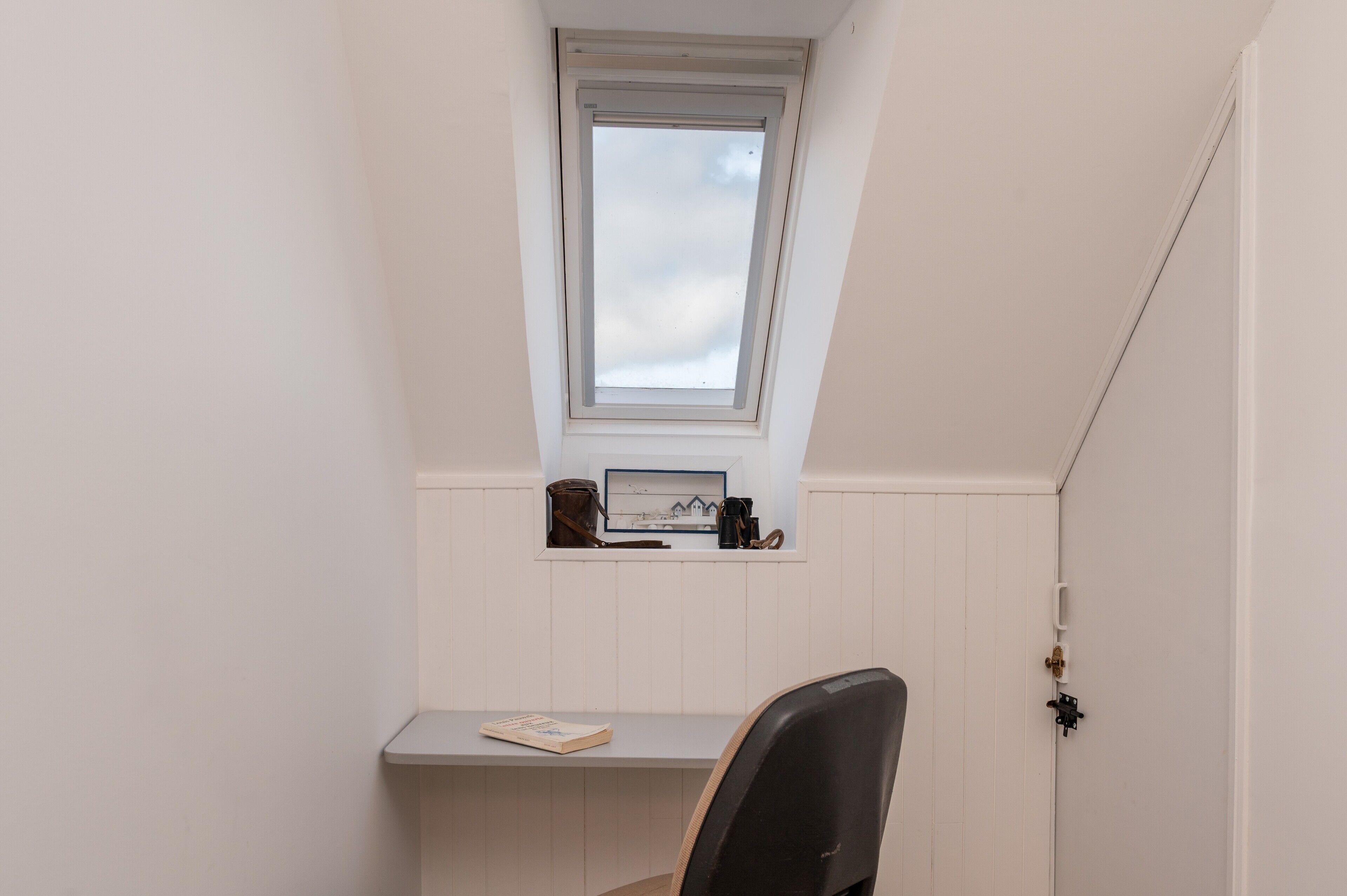 A room with a sloped ceiling, a small window, a desk with a chair, and a door.