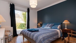 Una camera da letto con un letto, due comodini con lampade, una sedia, una finestra con tende e vista sul verde.