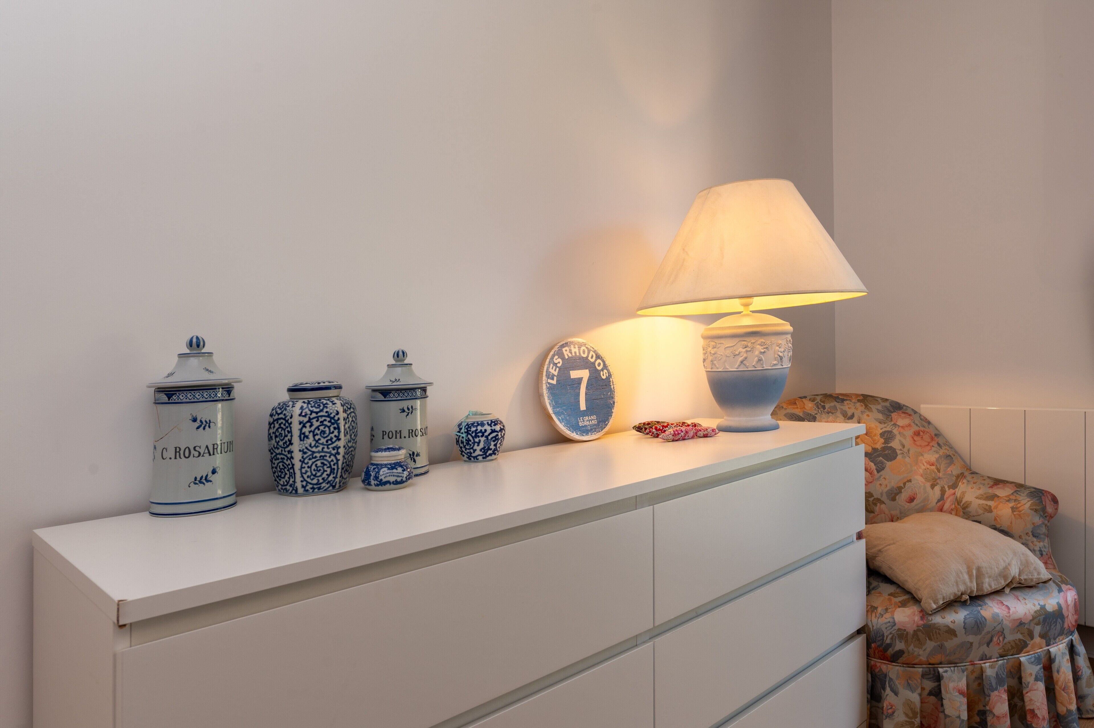 Een witte nachtkastje met een lamp, een klok en keramieken potjes.
