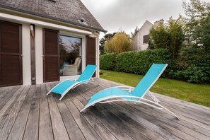 Terraza de madera con dos sillones reclinables y un ventanal de vidrio que da a una habitación.