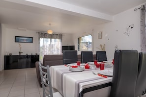 Un coin repas avec une table dressée pour quatre personnes, des chaises, un canapé noir, un téléviseur et une fenêtre avec des rideaux.