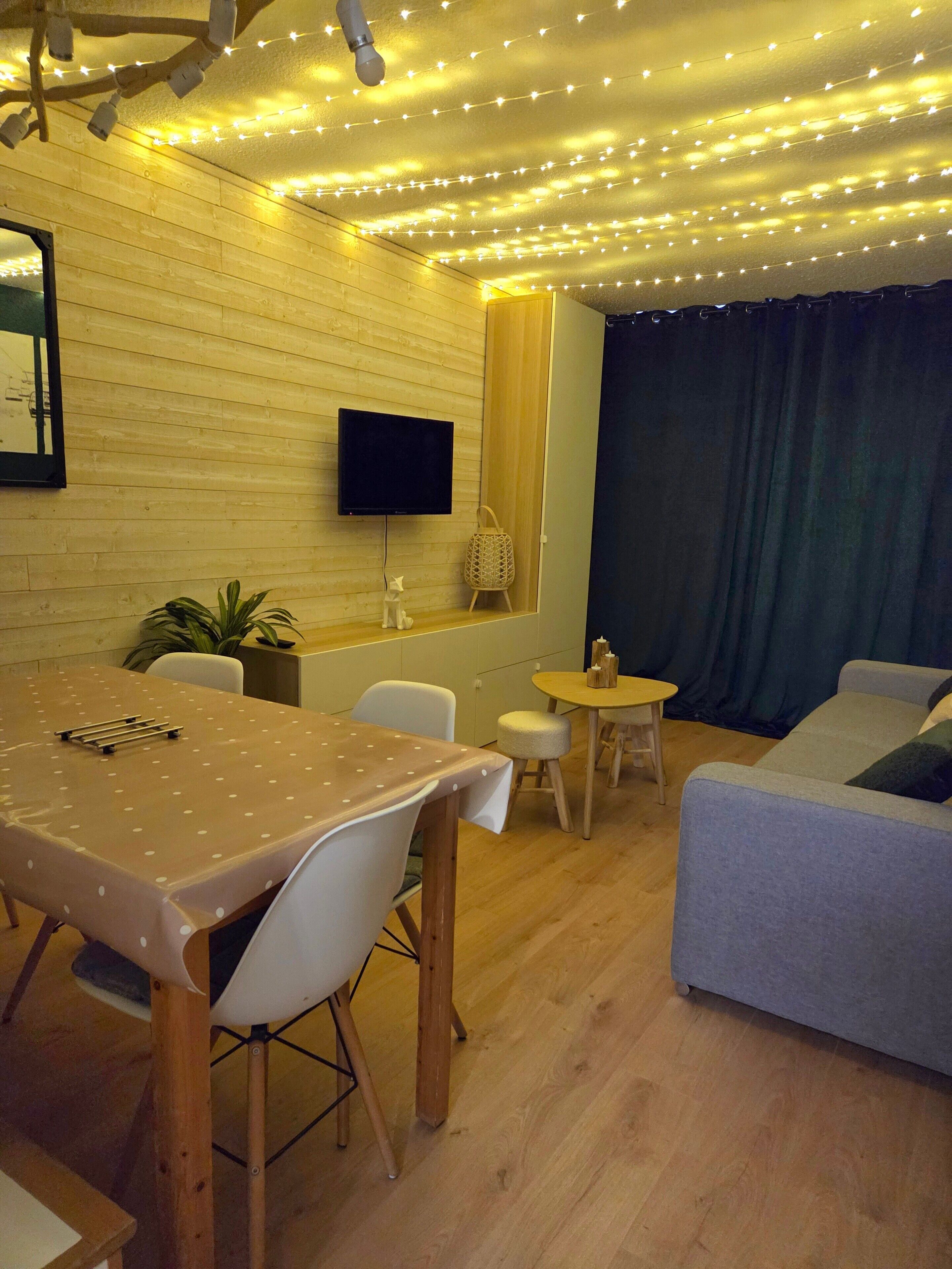 A cozy living room with a wooden floor, a dining table with chairs, a sofa, a small round table, and a TV mounted on the wall.