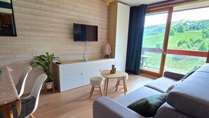Una sala de estar moderna con sofá, mesa de comedor y sillas, un televisor de pantalla plana montado en la pared y un ventanal con vistas a la vegetación.