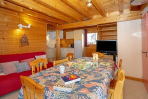 A cozy cabin interior with a wooden ceiling, a dining table with chairs, a sofa, a television, and a kitchen area.