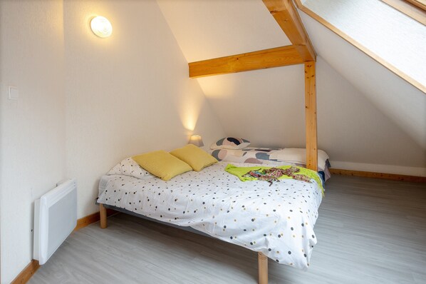 A bedroom with a sloped ceiling, a bed with a polka dot bedspread, two pillows, a nightstand with a lamp, and a radiator.