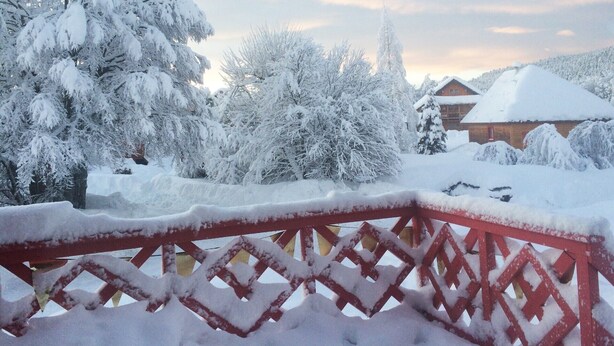 A snow-covered landscape with a red metal fence, trees, and a building in the background.