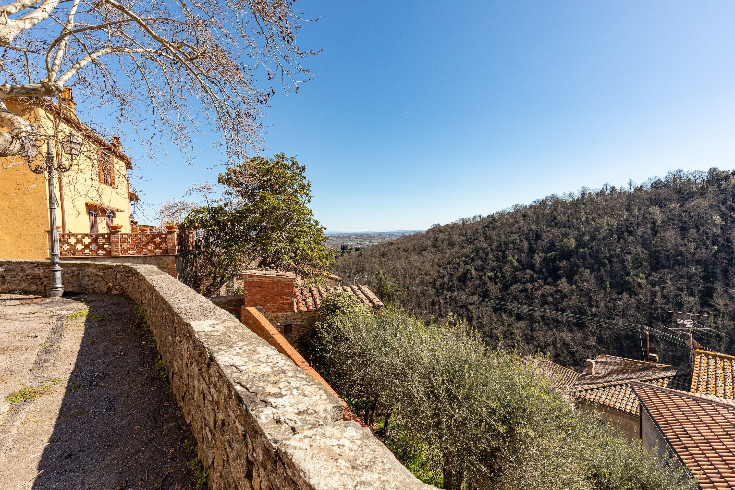 A hillside building with a stone wall, a view of the valley, and a forested hill in the background.