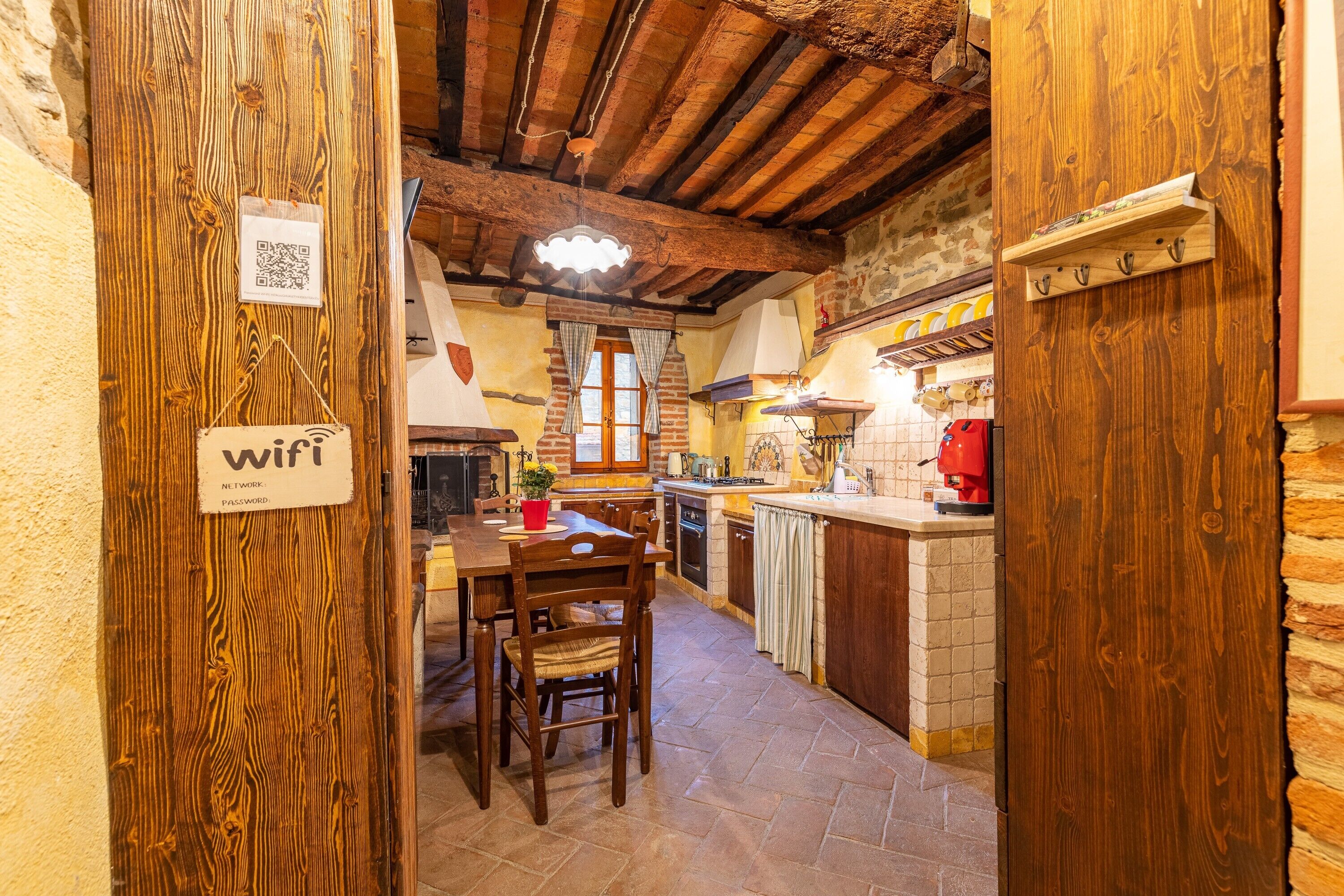 A rustic kitchen with exposed wooden beams, a dining table with chairs, a hanging light fixture, and a QR code for WiFi access.