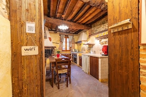 Una cocina rústica con vigas de madera a la vista, una mesa de comedor con sillas, una lámpara colgante y un código QR para acceso a WiFi.