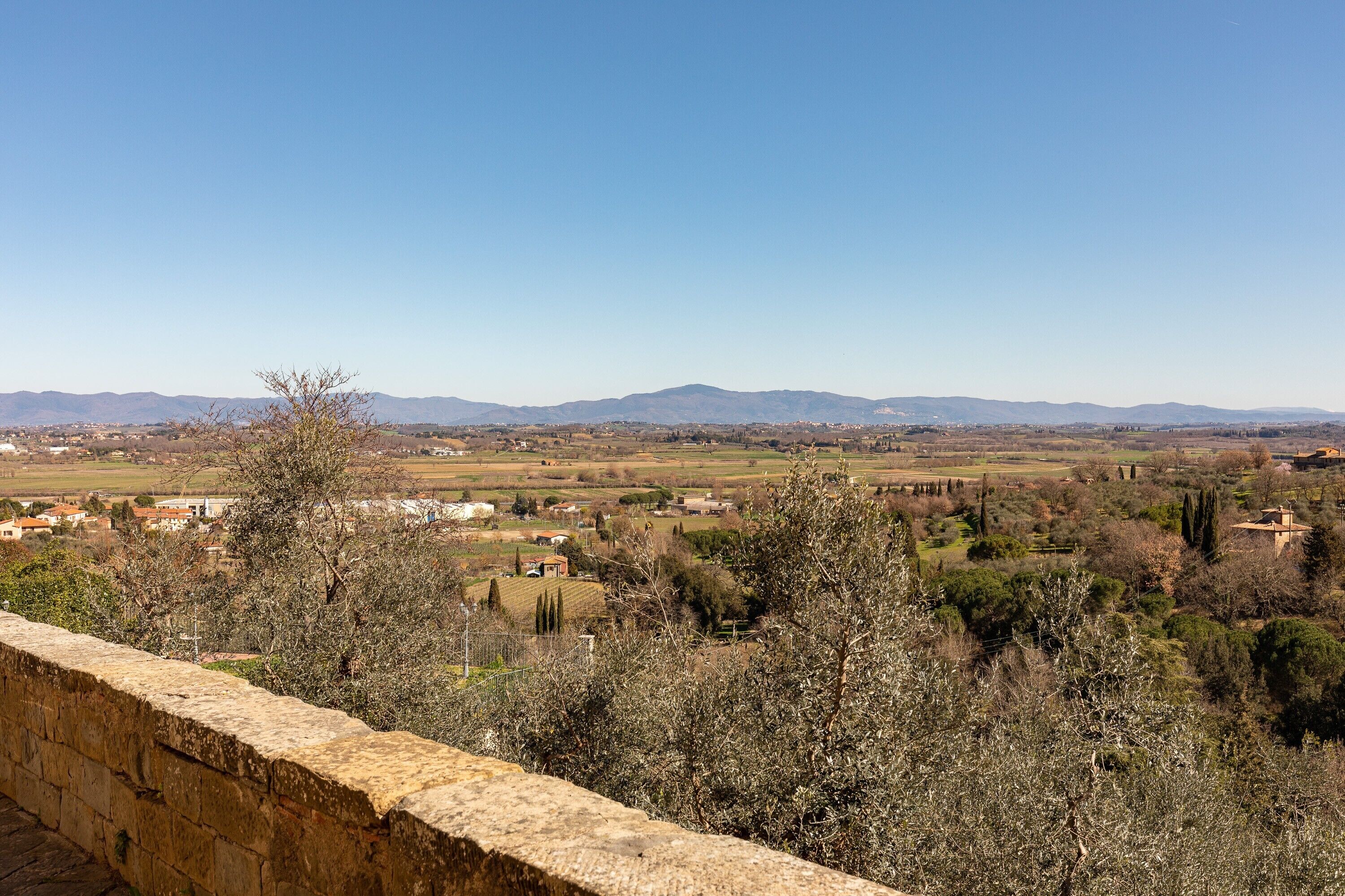 Mur de pedra amb vista a un paisatge rural, incloent camps, arbres i muntanyes allunyades.