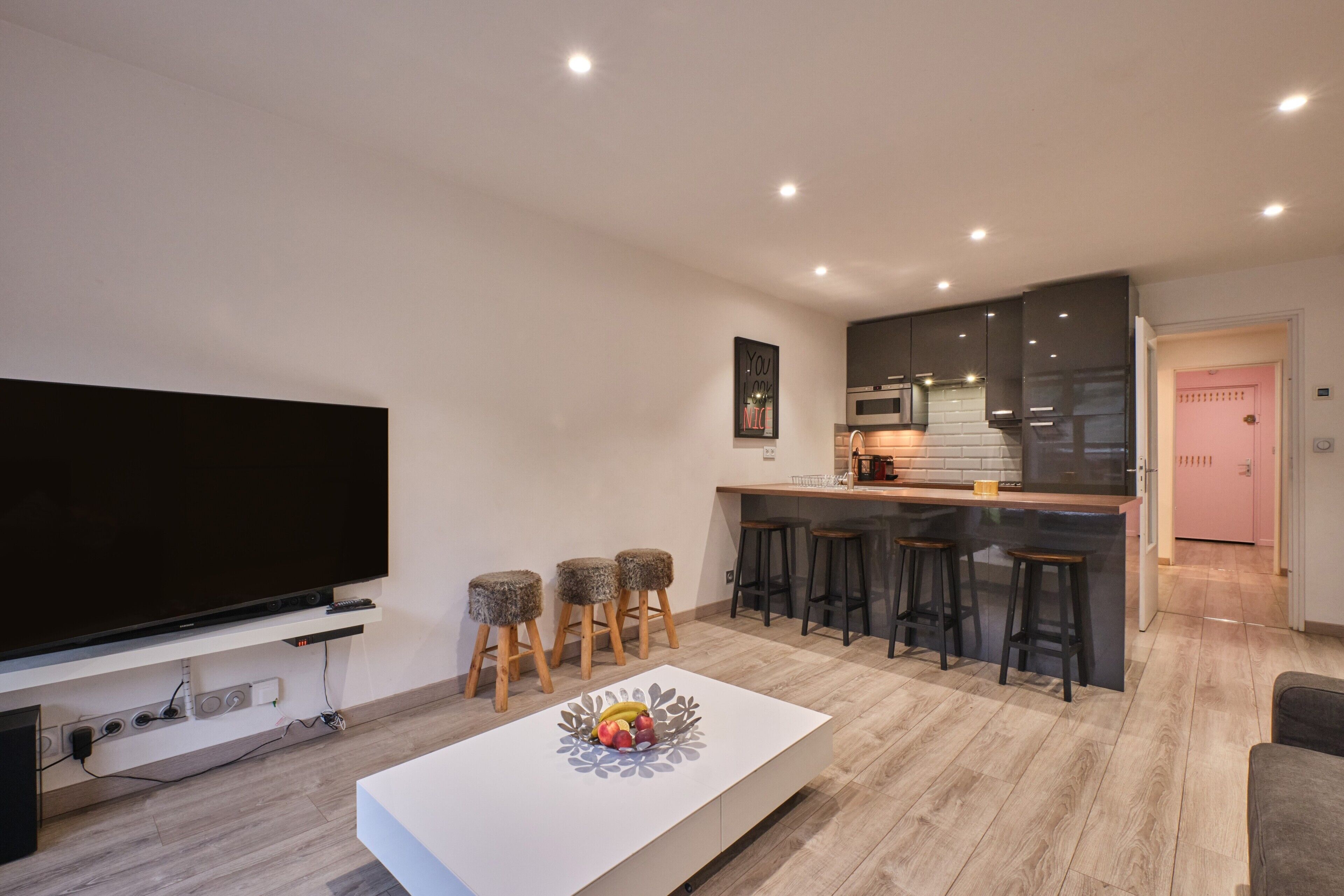 A modern living room with a flat-screen TV, a white coffee table with fruit, a kitchen area with bar stools, and a pink door.
