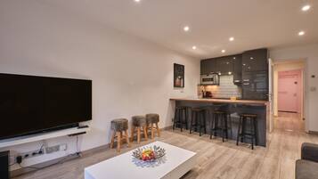 A modern living room with a flat-screen TV, a white coffee table with fruit, a kitchen area with bar stools, and a pink door.