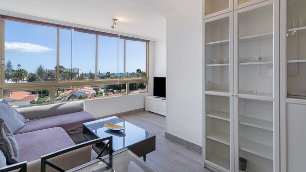 A modern living room with a sofa, a glass coffee table, a TV, and a large window offering a view of buildings and trees.