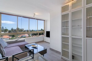 A modern living room with a sofa, a glass coffee table, a TV, and a large window offering a view of buildings and trees.