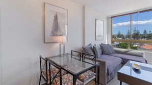 A modern living room with a glass dining table, a sofa with patterned cushions, and a large window offering a view of trees and buildings.