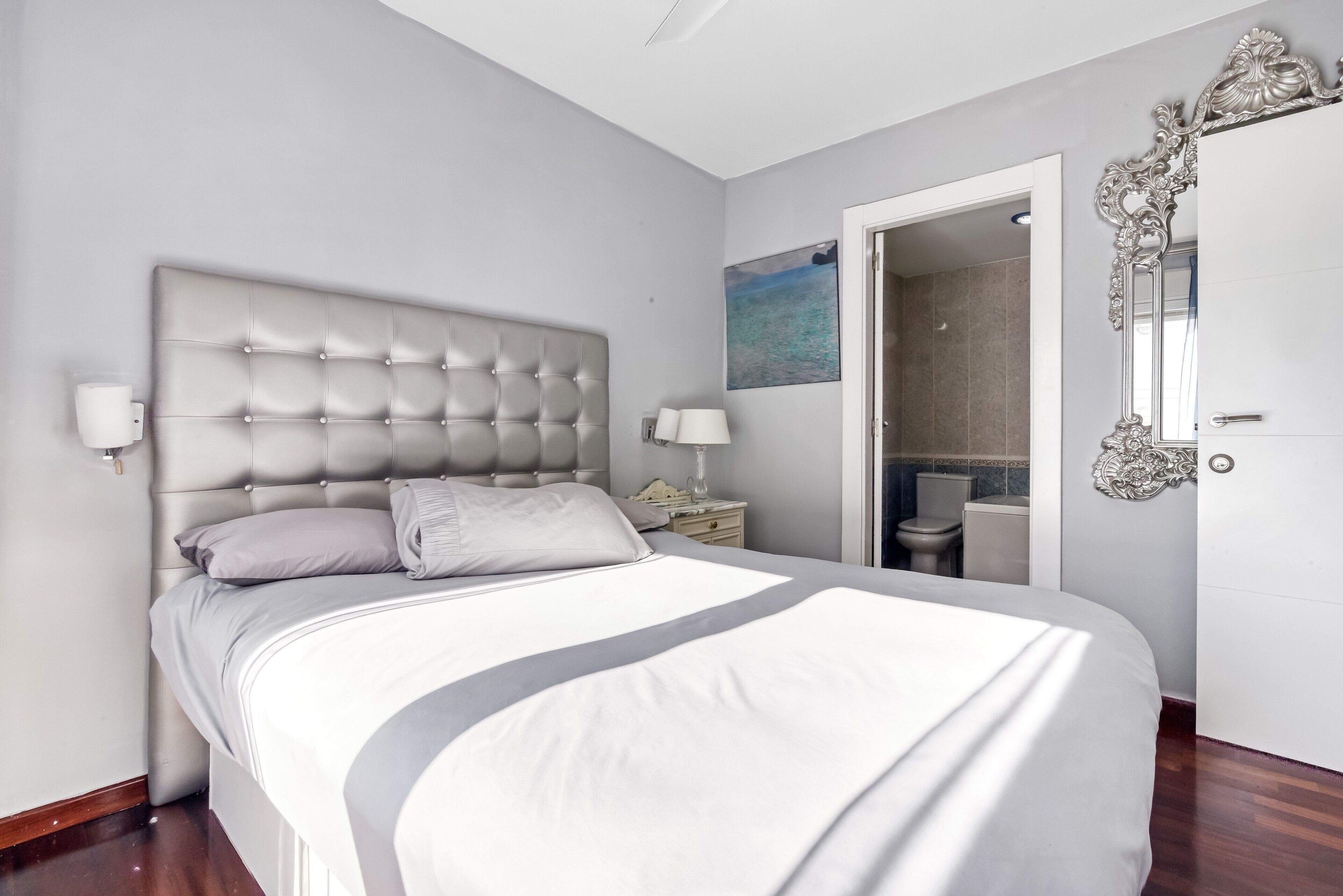 A bedroom with a tufted headboard, a bed with white bedding, a nightstand with a lamp, a bathroom door, and a decorative mirror.