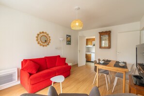 Een woonkamer met een rode bank, een eettafel met stoelen, een plafondlamp en een keuken die zichtbaar is door een open deur.