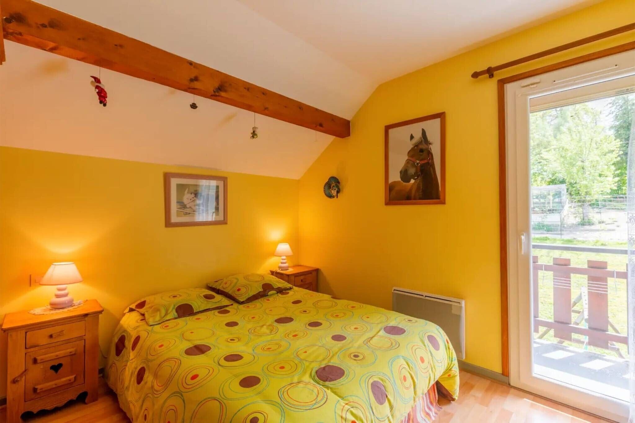 Une chambre à coucher avec un lit, deux tables de chevet avec des lampes, un radiateur, une poutre en bois au plafond, un tableau représentant un cheval et une porte-fenêtre coulissante donnant sur l’extérieur.