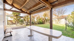Un patio cubierto con una mesa y bancos blancos, rodeado de un jardín y vistas a una zona residencial.