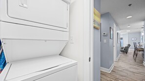 A hallway with a white appliance, a dining area with chairs and a table, and a wall with a clock and a thermostat.